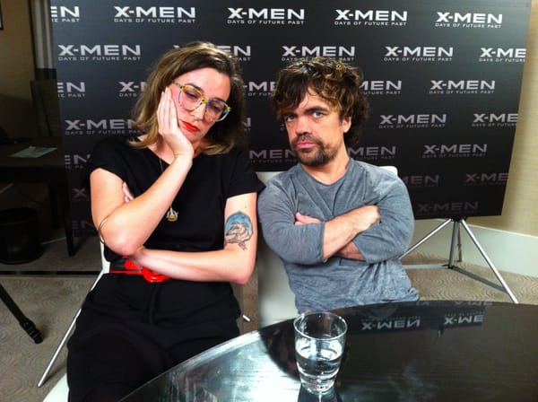 Clem, a white person in a black dress with a red belt, sits next to Peter Dinklage, a white guy wearing a grey marle long-sleeve. They're pretending to be bored in each other's company.